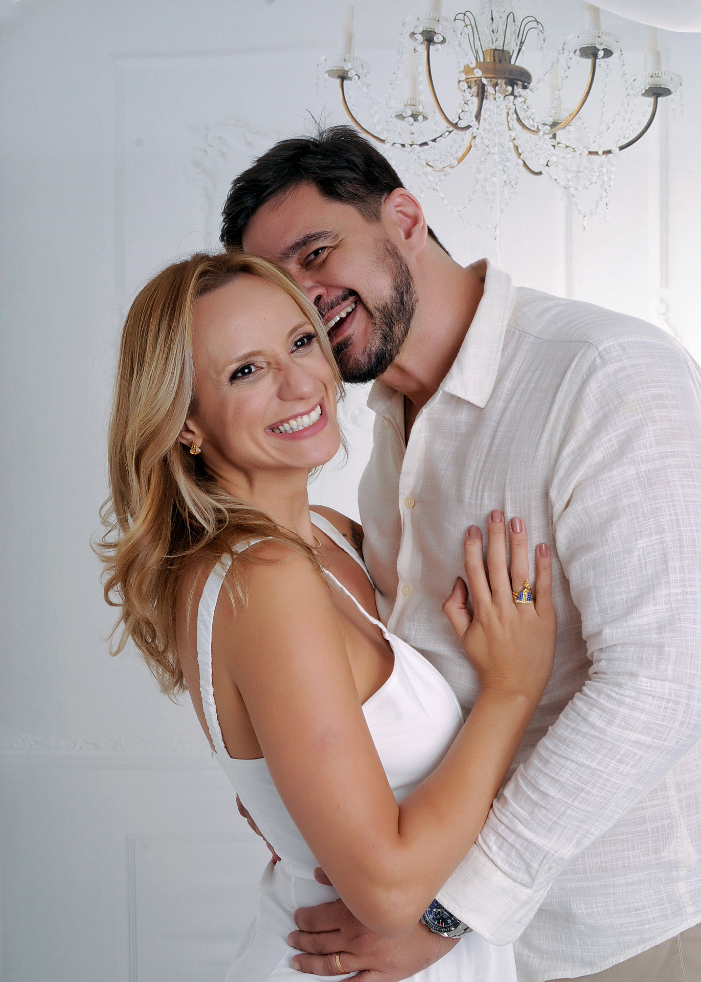 Fernanda Zago e o marido. Ela de branco sorrindo olhando para a câmera, ele por trás abraçando, fundo branco com lustre.